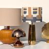 Une sélection de lampes design sur une étagère, avec la lampe à poser en grès ocre, la lampe champignon cuivrée, la lampe dorée à l'abat-jour graphique, et la lampe à poser gingko basse (un modèle haut est aussi proposé).