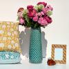 Objets déco de la collection Deco-Delight : des coussins d'été colorés, un case bleu en céramique, un cadre photo doré Art Déco, un sulfure oiseau ambré (statuette en verre soufflé), des fleurs artificielles (pivoines roses et allium).